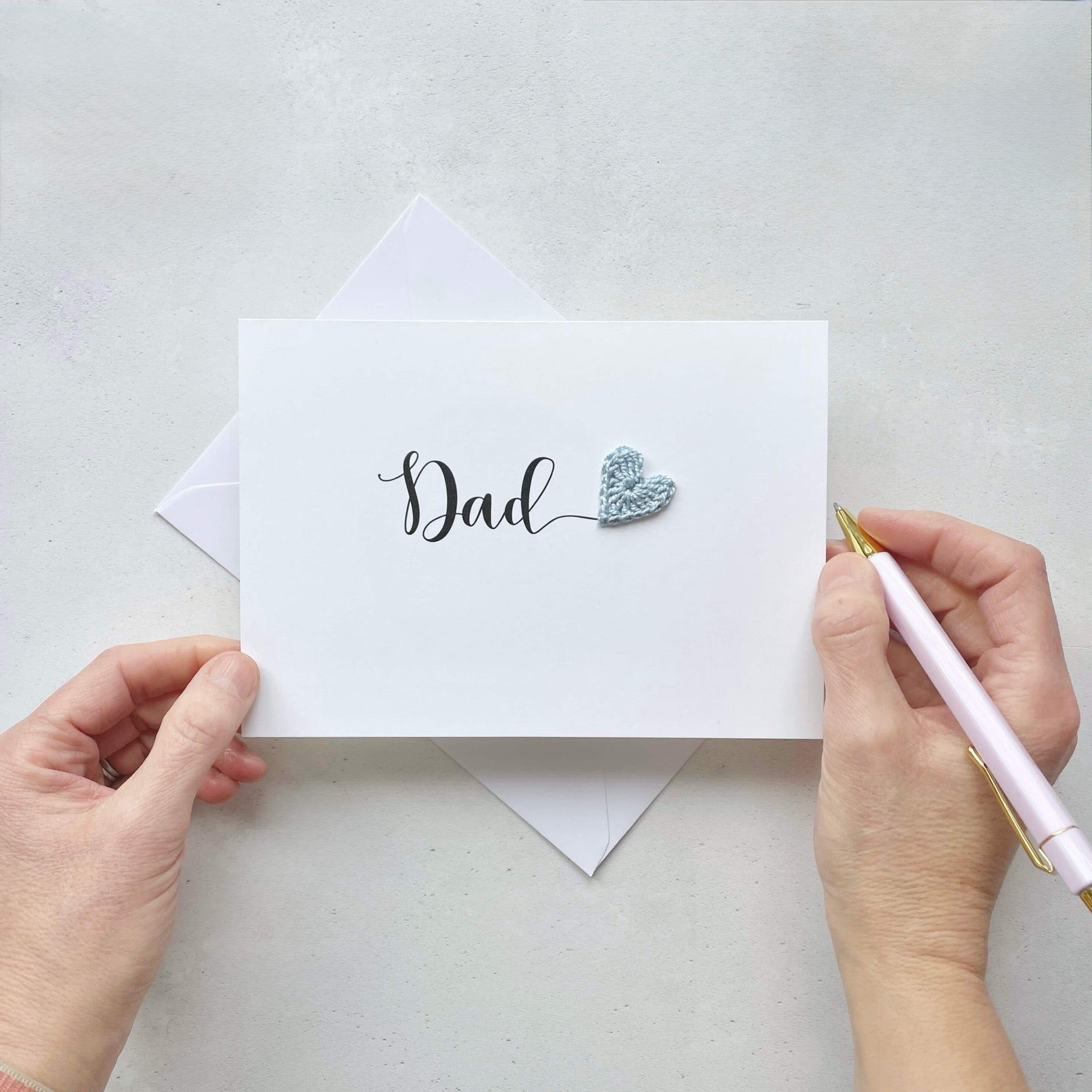 A white card with the word 'Dad' printed across in large black cursive text. A pale blue crochet heart has been placed at the end of the word. adding a pop of colour. 