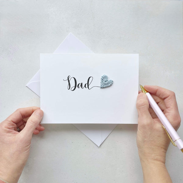 A white card with the word 'Dad' printed across in large black cursive text. A pale blue crochet heart has been placed at the end of the word. adding a pop of colour. 