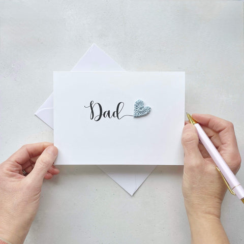 A white card with the word 'Dad' printed across in large black cursive text. A pale blue crochet heart has been placed at the end of the word. adding a pop of colour. 