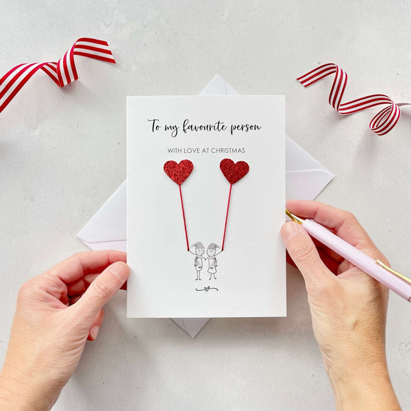 A Christmas card featuring a minimalist design with two figures wearing santa hats holding red glitter heart balloons. The text at the top reads: ‘To my favourite person, withh love at Christmas’. The card is placed on a white background with a white envelope partially visible behind it. A hand is seen holding the card, while another hand holds a pink pen. A red and white striped ribbon is positioned in the top corners of the image for decoration.