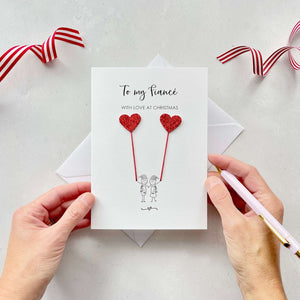 A Christmas card featuring a minimalist design with two figures wearing santa hats holding red glitter heart balloons. The text at the top reads: "To my Fiancé, With Love at Christmas." The card is placed on a white background with a white envelope partially visible behind it. A hand is seen holding the card, while another hand holds a pink pen. A red and white striped ribbon is positioned in the top corners of the image for decoration.