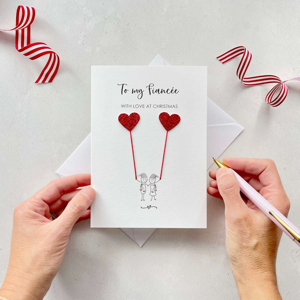 A Christmas card featuring a minimalist design with two figures wearing santa hats holding red glitter heart balloons. The text at the top reads: "To my Fiancée, With Love at Christmas." The card is placed on a white background with a white envelope partially visible behind it. A hand is seen holding the card, while another hand holds a pink pen. A red and white striped ribbon is positioned in the top corners of the image for decoration.