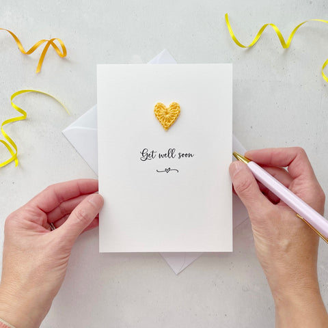 A white portrait-style card featuring the word ‘Get well soon’ in elegant black cursive at the center. Below the text, a small black squiggle and heart design adds a playful touch. Above the word, a handmade yellow crochet heart is carefully attached, providing a pop of colour and texture.