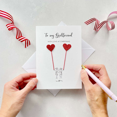 A Christmas card featuring a minimalist design with two figures wearing santa hats holding red glitter heart balloons. The text at the top reads: ‘To my Girlfriend, with love at Christmas’. The card is placed on a white background with a white envelope partially visible behind it. A hand is seen holding the card, while another hand holds a pink pen. A red and white striped ribbon is positioned in the top corners of the image for decoration.