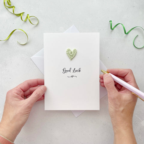 A white portrait-style card featuring the word ‘Good luck’ in elegant black cursive at the center. Below the text, a small black squiggle and heart design adds a playful touch. Above the word, a handmade pale green crochet heart is carefully attached, providing a pop of colour and texture.