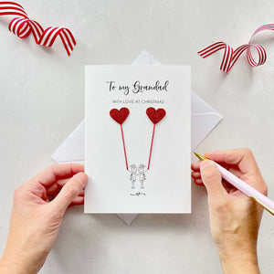 A Christmas card featuring a minimalist design with two figures wearing santa hats holding red glitter heart balloons. The text at the top reads: "To my Grandad, With Love at Christmas." The card is placed on a white background with a white envelope partially visible behind it. A hand is seen holding the card, while another hand holds a pink pen. A red and white striped ribbon is positioned in the top corners of the image for decoration.