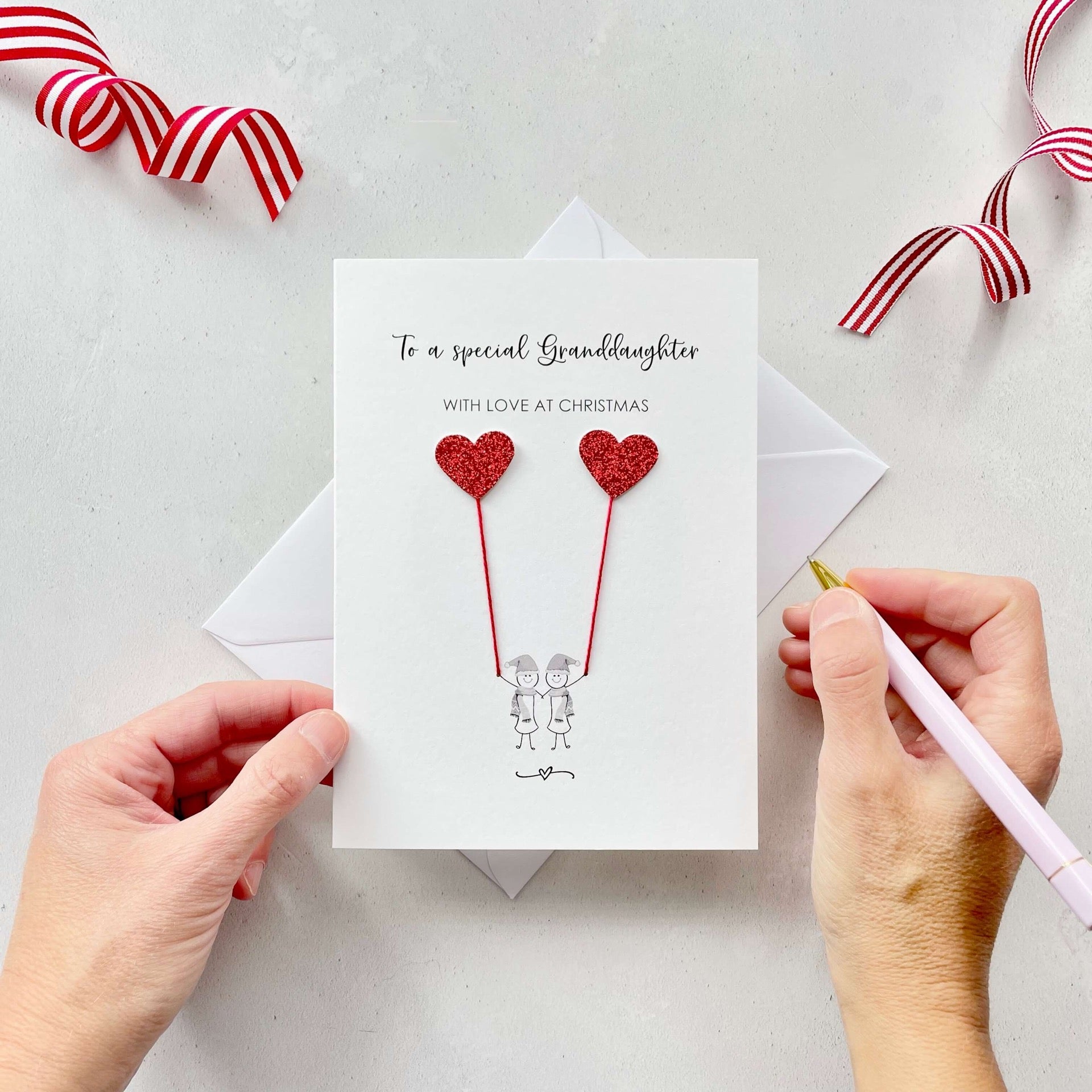 A Christmas card featuring a minimalist design with two figures wearing santa hats holding red glitter heart balloons. The text at the top reads: "To a special Granddaughter, With Love at Christmas." The card is placed on a white background with a white envelope partially visible behind it. A hand is seen holding the card, while another hand holds a pink pen. A red and white striped ribbon is positioned in the top corners of the image for decoration.