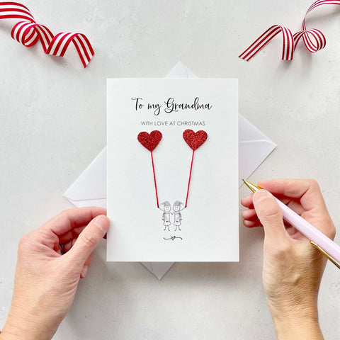 A Christmas card featuring a minimalist design with two figures wearing santa hats holding red glitter heart balloons. The text at the top reads: "To my Grandma, With Love at Christmas." The card is placed on a white background with a white envelope partially visible behind it. A hand is seen holding the card, while another hand holds a pink pen. A red and white striped ribbon is positioned in the top corners of the image for decoration.