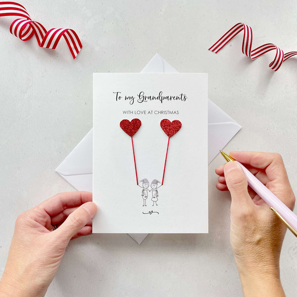 A Christmas card featuring a minimalist design with two figures wearing santa hats holding red glitter heart balloons. The text at the top reads: "To my Grandparents, With Love at Christmas." The card is placed on a white background with a white envelope partially visible behind it. A hand is seen holding the card, while another hand holds a pink pen. A red and white striped ribbon is positioned in the top corners of the image for decoration.