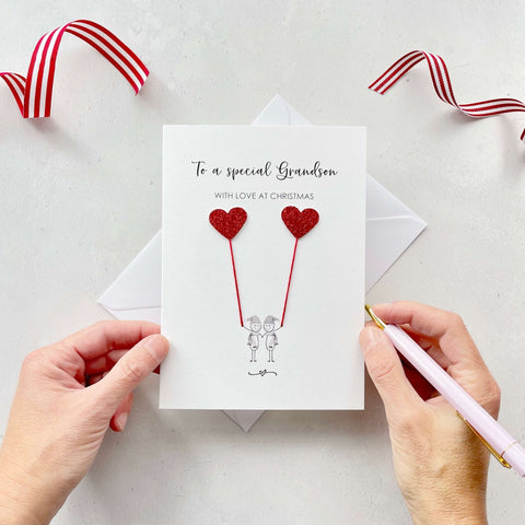 A Christmas card featuring a minimalist design with two figures wearing santa hats holding red glitter heart balloons. The text at the top reads: "To a special Grandson, With Love at Christmas." The card is placed on a white background with a white envelope partially visible behind it. A hand is seen holding the card, while another hand holds a pink pen. A red and white striped ribbon is positioned in the top corners of the image for decoration.