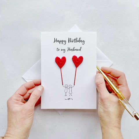 A white birthday card featuring a line drawn couple holding hands and each holding a red crochet heart balloon.  ‘Happy Birthday to my Husband’ is printed at the top of the card in black cursive text.