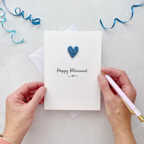 A white portrait-style card featuring the words ‘Happy Retirement’ in elegant black cursive at the center. Below the text, a small black squiggle and heart design adds a playful touch. Above the words, a handmade blue crochet heart is carefully attached, providing a pop of colour and texture.