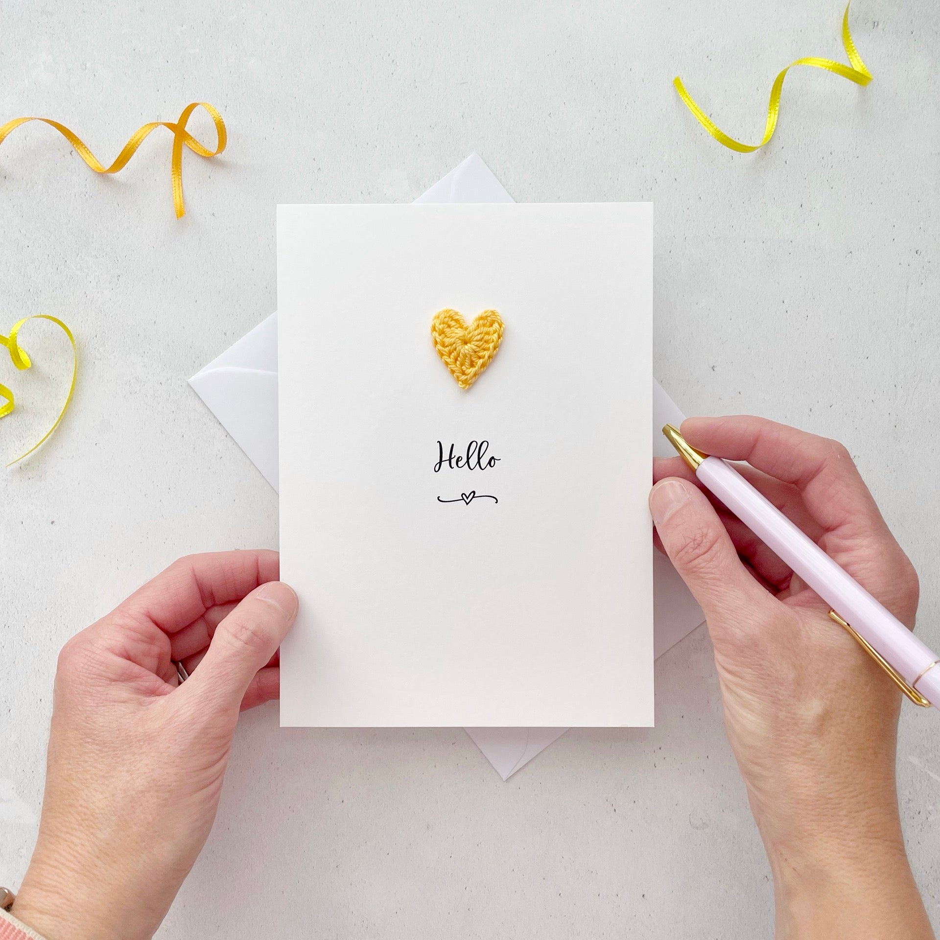 A white portrait-style card featuring the word ‘Hello’ in elegant black cursive at the center. Below the text, a small black squiggle and heart design adds a playful touch. Above the word, a handmade yellow crochet heart is carefully attached, providing a pop of color and texture.