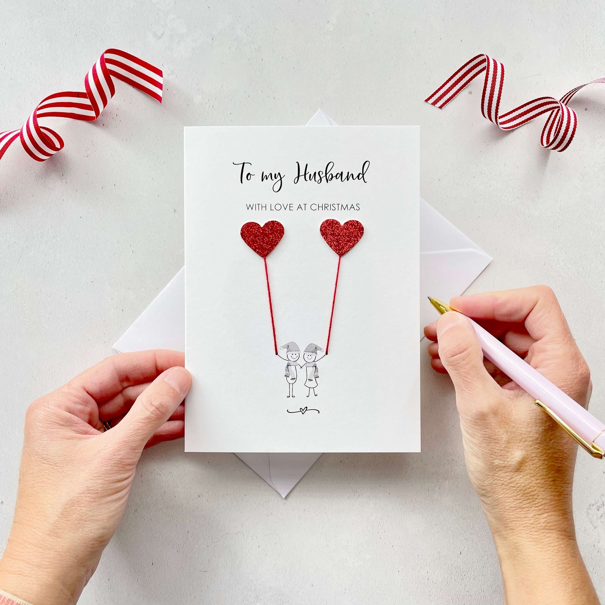 A Christmas card featuring a minimalist design with two figures wearing santa hats holding red glitter heart balloons. The text at the top reads: "To my Husband, With Love at Christmas." The card is placed on a white background with a white envelope partially visible behind it. A hand is seen holding the card, while another hand holds a pink pen. A red and white striped ribbon is positioned in the top corners of the image for decoration.