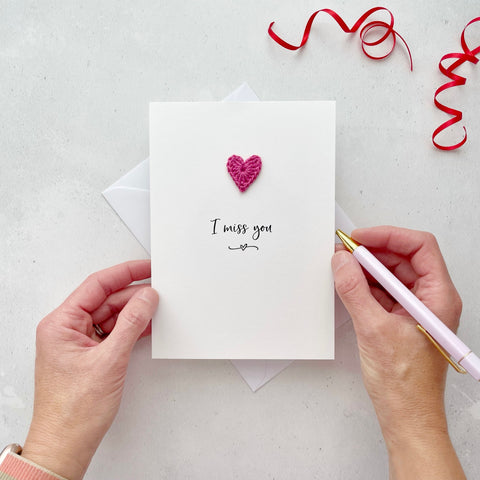 A white portrait-style card featuring the words ‘I miss you’ in elegant black cursive at the center. Below the text, a small black squiggle and heart design adds a playful touch. Above the words, a handmade bright pink crochet heart is carefully attached, providing a pop of colour and texture.