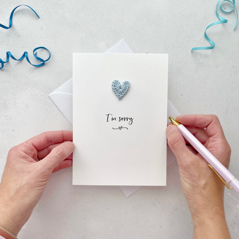 A white portrait-style card featuring the words ‘I’m sorry’ in elegant black cursive at the center. Below the text, a small black squiggle and heart design adds a playful touch. Above the words, a handmade pale blue crochet heart is carefully attached, providing a pop of color and texture.