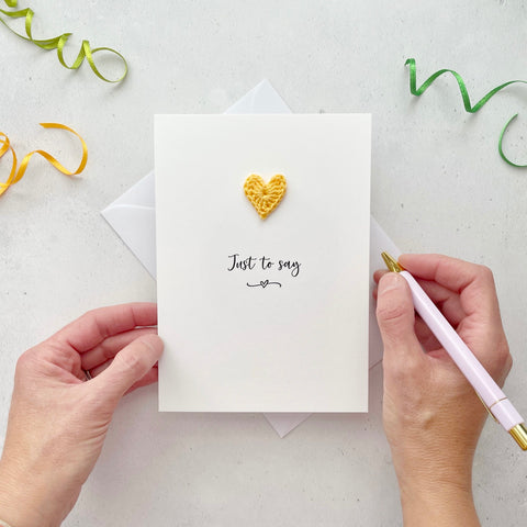 A white portrait-style card featuring the words ‘Just to say’ in elegant black cursive at the center. Below the text, a small black squiggle and heart design adds a playful touch. Above the words, a handmade yellow crochet heart is carefully attached, providing a pop of colour and texture.