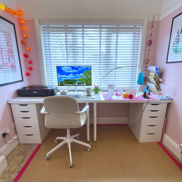 Mollycat Studio work space. A long white desk situated below a window. The walls are light pink and there are prints on the wall along with colourful festoon lights. 