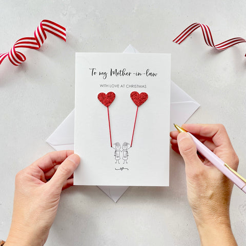 A Christmas card featuring a minimalist design with two figures wearing santa hats holding red glitter heart balloons. The text at the top reads: ‘To my Mother in law, with love at Christmas’. The card is placed on a white background with a white envelope partially visible behind it. A hand is seen holding the card, while another hand holds a pink pen. A red and white striped ribbon is positioned in the top corners of the image for decoration.