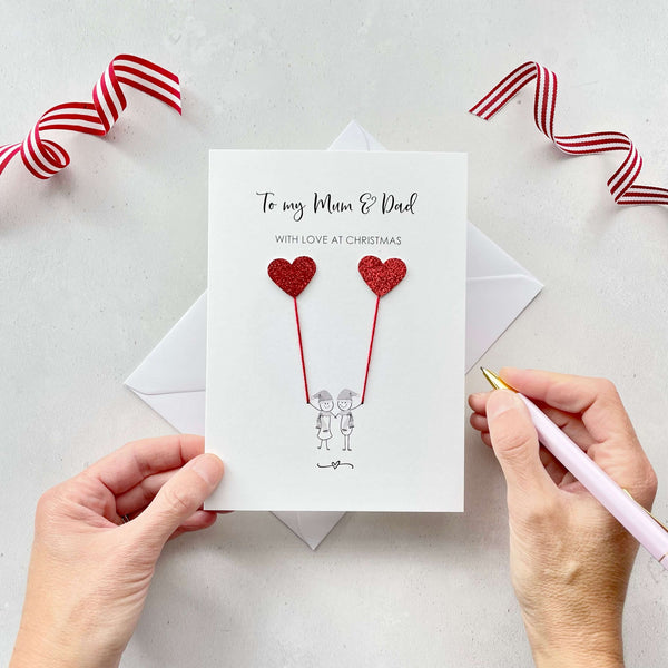 A Christmas card featuring a minimalist design with two figures wearing santa hats holding red glitter heart balloons. The text at the top reads: "To my Mum & Dad, With Love at Christmas." The card is placed on a white background with a white envelope partially visible behind it. A hand is seen holding the card, while another hand holds a pink pen. A red and white striped ribbon is positioned in the top corners of the image for decoration.