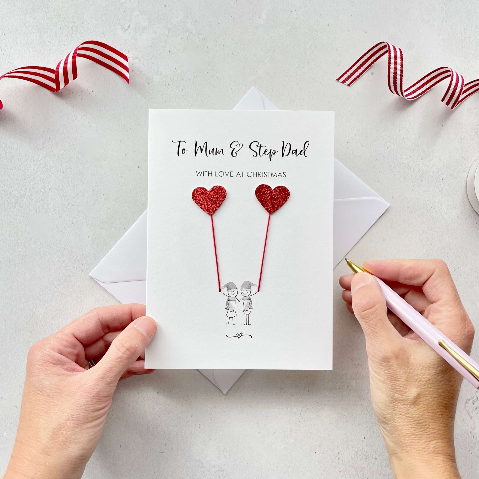 A Christmas card featuring a minimalist design with two figures wearing santa hats holding red glitter heart balloons. The text at the top reads: ‘To Mum & Step Dad, with love at Christmas’. The card is placed on a white background with a white envelope partially visible behind it. A hand is seen holding the card, while another hand holds a pink pen. A red and white striped ribbon is positioned in the top corners of the image for decoration.