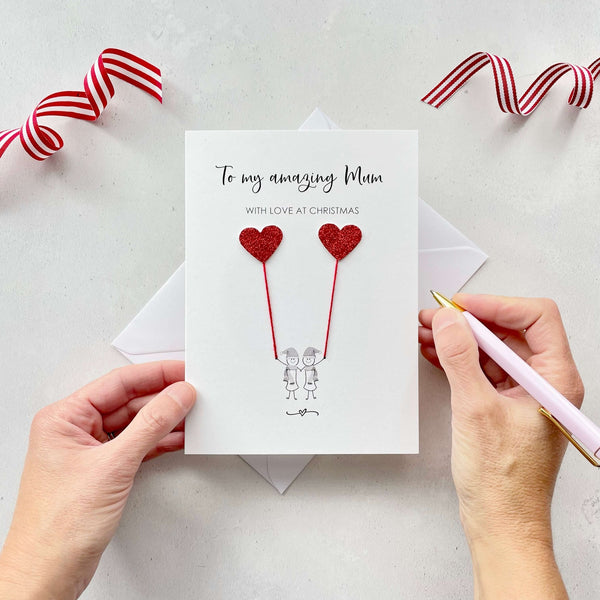 A Christmas card featuring a minimalist design with two figures wearing santa hats holding red glitter heart balloons. The text at the top reads: "To my amazing Mum, With Love at Christmas." The card is placed on a white background with a white envelope partially visible behind it. A hand is seen holding the card, while another hand holds a pink pen. A red and white striped ribbon is positioned in the top corners of the image for decoration.