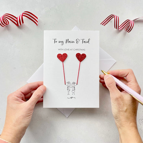 A Christmas card featuring a minimalist design with two figures wearing santa hats holding red glitter heart balloons. The text at the top reads: ‘To my Nain & Taid, With Love at Christmas’. The card is placed on a white background with a white envelope partially visible behind it. A hand is seen holding the card, while another hand holds a pink pen. A red and white striped ribbon is positioned in the top corners of the image for decoration.
