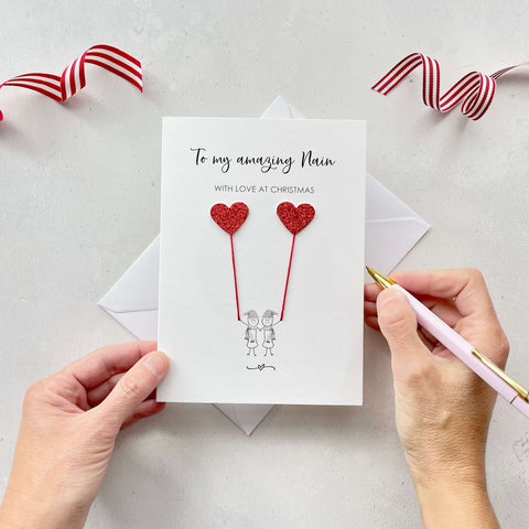 A Christmas card featuring a minimalist design with two figures wearing santa hats holding red glitter heart balloons. The text at the top reads: ‘To my amazing Nain, With Love at Christmas’. The card is placed on a white background with a white envelope partially visible behind it. A hand is seen holding the card, while another hand holds a pink pen. A red and white striped ribbon is positioned in the top corners of the image for decoration.