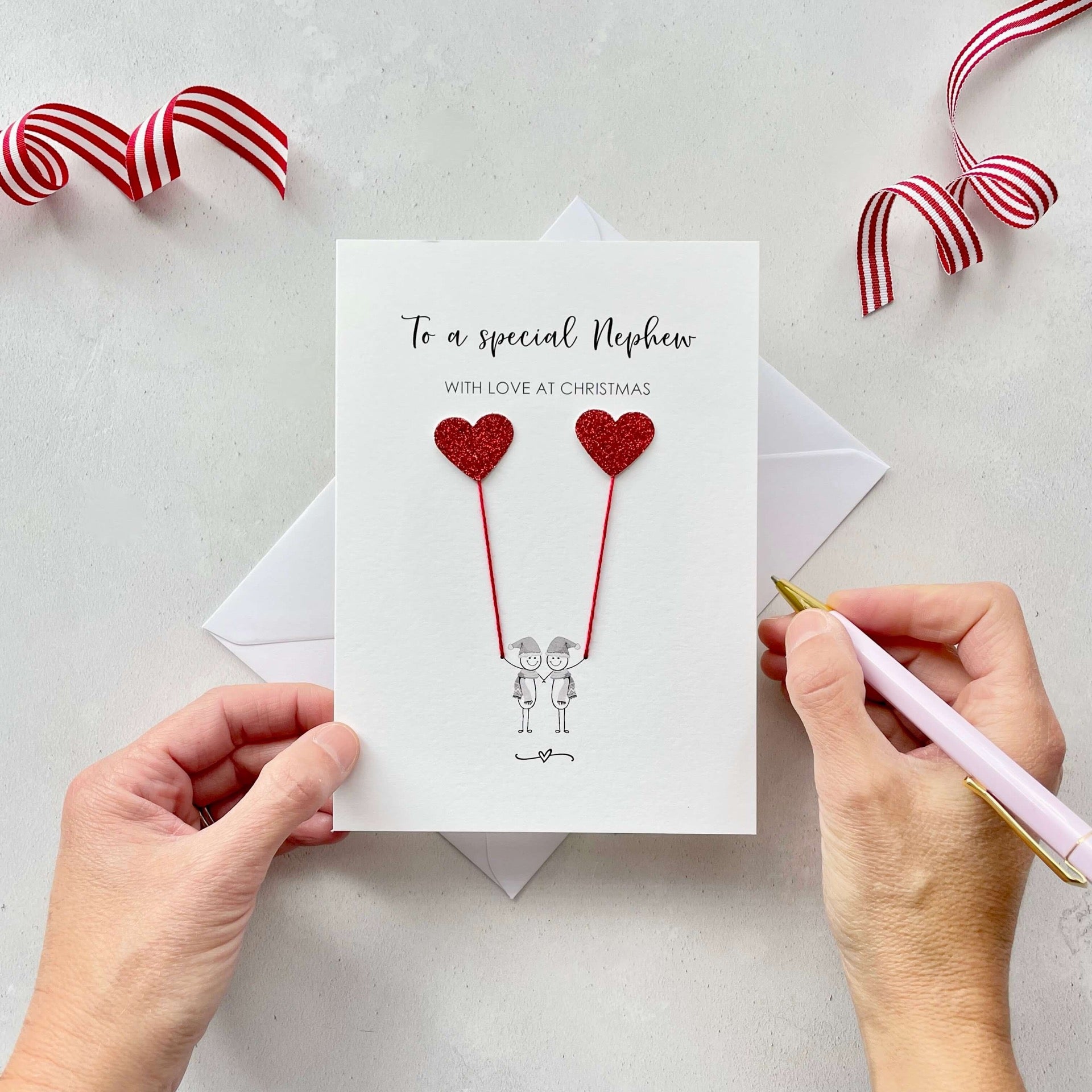 A Christmas card featuring a minimalist design with two figures wearing santa hats holding red glitter heart balloons. The text at the top reads: ‘To a special Nephew, with love at Christmas’. The card is placed on a white background with a white envelope partially visible behind it. A hand is seen holding the card, while another hand holds a pink pen. A red and white striped ribbon is positioned in the top corners of the image for decoration.