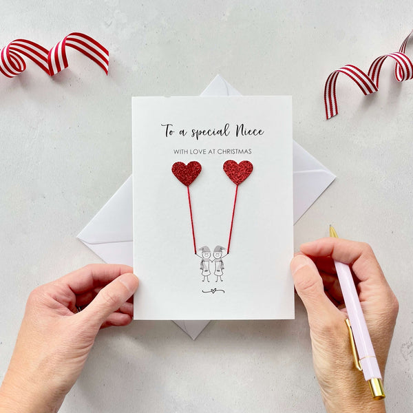 A Christmas card featuring a minimalist design with two figures wearing santa hats holding red glitter heart balloons. The text at the top reads: ‘To a special Niece, with love at Christmas’. The card is placed on a white background with a white envelope partially visible behind it. A hand is seen holding the card, while another hand holds a pink pen. A red and white striped ribbon is positioned in the top corners of the image for decoration.