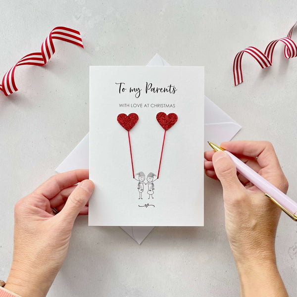 A Christmas card featuring a minimalist design with two figures wearing santa hats holding red glitter heart balloons. The text at the top reads: ‘To my parents, with love at Christmas’. The card is placed on a white background with a white envelope partially visible behind it. A hand is seen holding the card, while another hand holds a pink pen. A red and white striped ribbon is positioned in the top corners of the image for decoration.