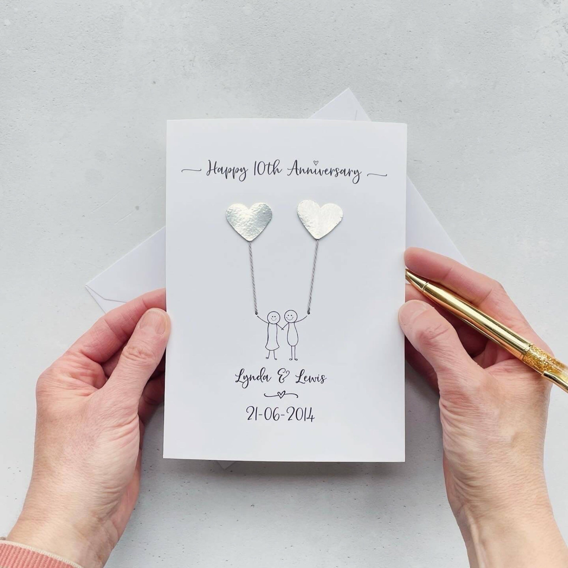 The image shows a beautiful handmade anniversary card with two silver mirror card hearts attached by grey yarn, resembling balloons being held by a hand-drawn couple underneath. The text on the card reads "Happy 10th Anniversary" at the top, followed by the couples names and wedding date at the bottom. The card is set on a light background, and the person's hands are seen holding a pen and gently holding the card, giving the image a sense of care and personalisation.