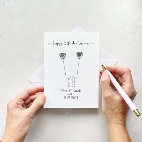 Handmade anniversary card embelished with 2 heart shaped plastic gems, both attached by cotton yarn, resembling balloons being held by a couple underneath. Happy 15th Anniversary is printed at the top, the couples names/ wedding date at the bottom.