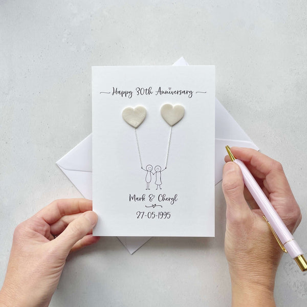 A portrait pearl anniversary card features two pearlised polymer clay hearts attached by cotton yarn, creating the illusion of balloons being held by a hand-drawn couple. The top of the card reads “Happy 30th Anniversary,” while the names and wedding date are printed at the bottom.