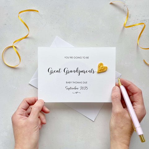 A landscape pregnancy announcement card with the text “You’re going to be Great Grandparents” in elegant black font. Below, it says “Baby Thomas due September 2025,” with a small decorative flourish. The card features a small crocheted yellow heart.