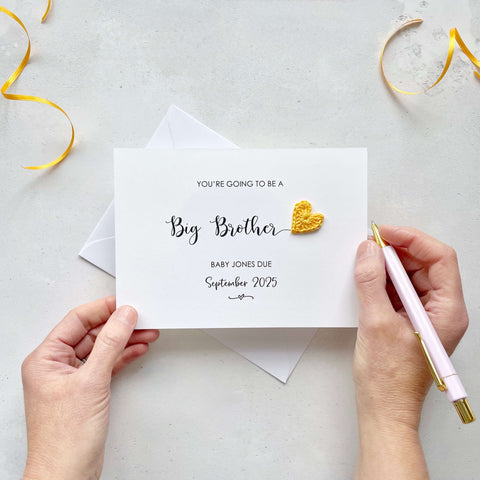 A landscape pregnancy announcement card with the text “You’re going to be a Big Brother” in elegant black font. Below, it says “Baby Jones due September 2025,” with a small decorative flourish. The card features a small crocheted yellow heart.