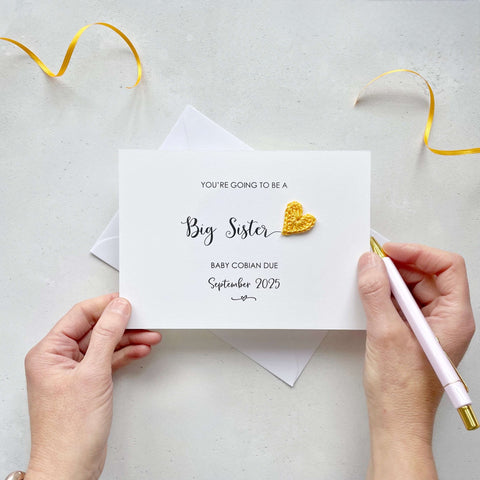 A landscape pregnancy announcement card with the text “You’re going to be a Big Sister” in elegant black font. Below, it says “Baby Cobain due September 2025,” with a small decorative flourish. The card features a small crocheted yellow heart.