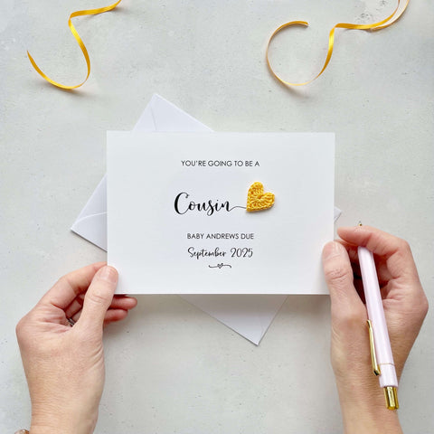 A landscape pregnancy announcement card with the text “You’re going to be a Cousin” in elegant black font. Below, it says “Baby Andrews due September 2025,” with a small decorative flourish. The card features a small crocheted yellow heart.