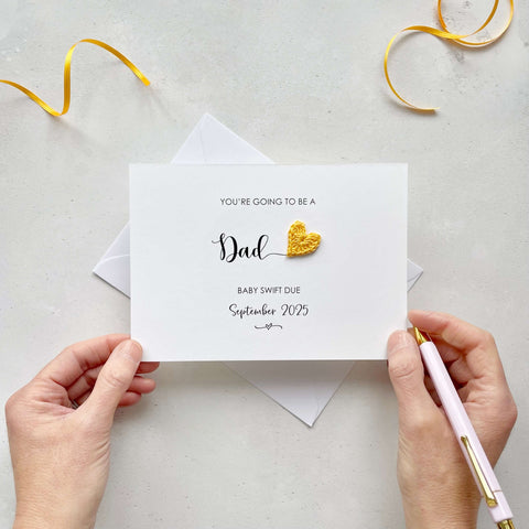 A landscape pregnancy announcement card with the text “You’re going to be a Dad” in elegant black font. Below, it says “Baby Swift due September 2025,” with a small decorative flourish. The card features a small crocheted yellow heart.