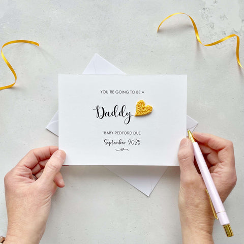 A landscape pregnancy announcement card with the text “You’re going to be a Daddy” in elegant black font. Below, it says “Baby Redford due September 2025,” with a small decorative flourish. The card features a small crocheted yellow heart.