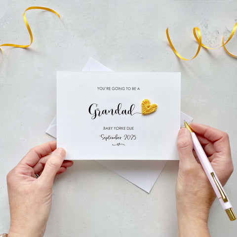 A landscape pregnancy announcement card with the text “You’re going to be a Grandad” in elegant black font. Below, it says “Baby Yorke due September 2025,” with a small decorative flourish. The card features a small crocheted yellow heart.