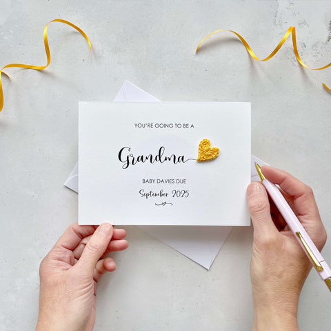 A landscape pregnancy announcement card with the text “You’re going to be a Grandma” in elegant black font. Below, it says “Baby Davies due September 2025,” with a small decorative flourish. The card features a small crocheted yellow heart.