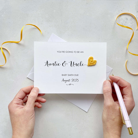 A landscape pregnancy announcement card with the text “You’re going to be an Auntie & Uncle” in elegant black font. Below, it says “Baby Smith due August 2025,” with a small decorative flourish. The card features a small crocheted yellow heart.