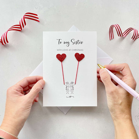 A Christmas card featuring a minimalist design with two figures wearing santa hats holding red glitter heart balloons. The text at the top reads: ‘To my Sister, With Love at Christmas’. The card is placed on a white background with a white envelope partially visible behind it. A hand is seen holding the card, while another hand holds a pink pen. A red and white striped ribbon is positioned in the top corners of the image for decoration.