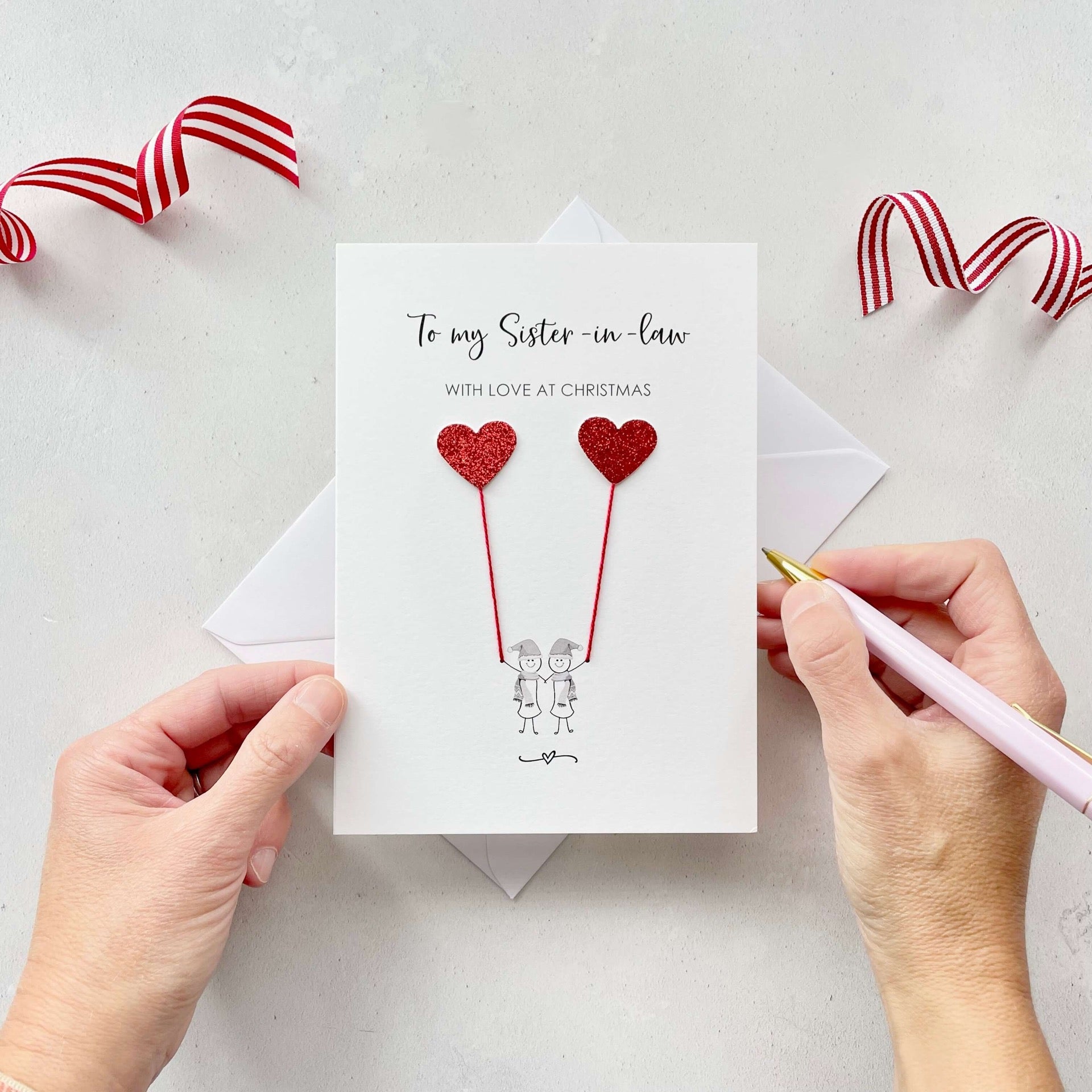 A Christmas card featuring a minimalist design with two figures wearing santa hats holding red glitter heart balloons. The text at the top reads: ‘To my Sister in law, with love at Christmas’. The card is placed on a white background with a white envelope partially visible behind it. A hand is seen holding the card, while another hand holds a pink pen. A red and white striped ribbon is positioned in the top corners of the image for decoration.