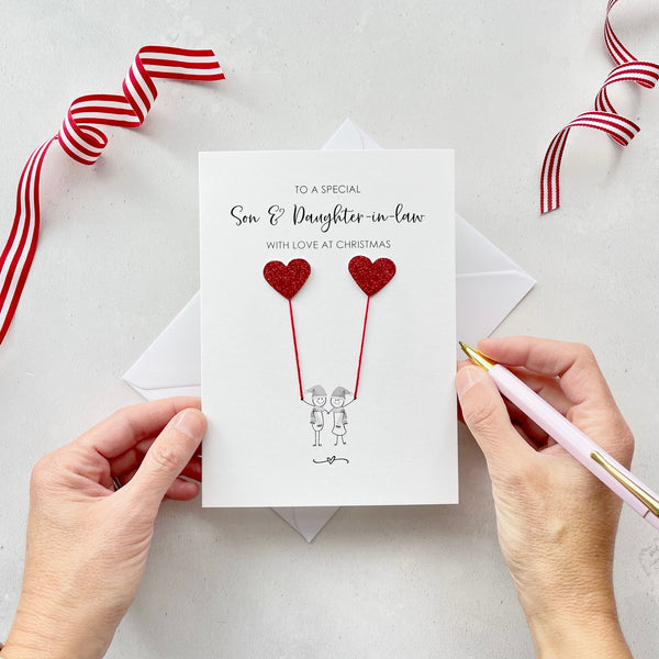 A Christmas card featuring a minimalist design with two figures wearing santa hats holding red glitter heart balloons. The text at the top reads: ‘To a special Son & Daughter-in-law, with love at Christmas’. The card is placed on a white background with a white envelope partially visible behind it. A hand is seen holding the card, while another hand holds a pink pen. A red and white striped ribbon is positioned in the top corners of the image for decoration.