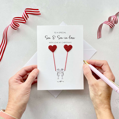 A Christmas card featuring a minimalist design with two figures wearing santa hats holding red glitter heart balloons. The text at the top reads: ‘To a special Son & Son-in-law, with love at Christmas’. The card is placed on a white background with a white envelope partially visible behind it. A hand is seen holding the card, while another hand holds a pink pen. A red and white striped ribbon is positioned in the top corners of the image for decoration.