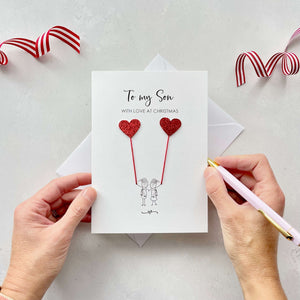 A Christmas card featuring a minimalist design with two figures wearing santa hats holding red glitter heart balloons. The text at the top reads: "To my Son, With Love at Christmas." The card is placed on a white background with a white envelope partially visible behind it. A hand is seen holding the card, while another hand holds a pink pen. A red and white striped ribbon is positioned in the top corners of the image for decoration.