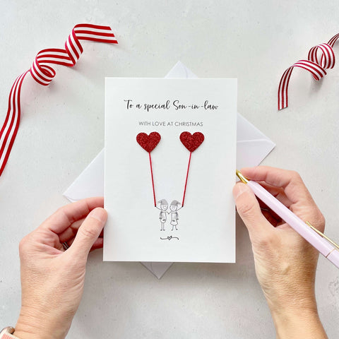 A Christmas card featuring a minimalist design with two figures wearing santa hats holding red glitter heart balloons. The text at the top reads: ‘To a special Son in law, with love at Christmas’. The card is placed on a white background with a white envelope partially visible behind it. A hand is seen holding the card, while another hand holds a pink pen. A red and white striped ribbon is positioned in the top corners of the image for decoration.