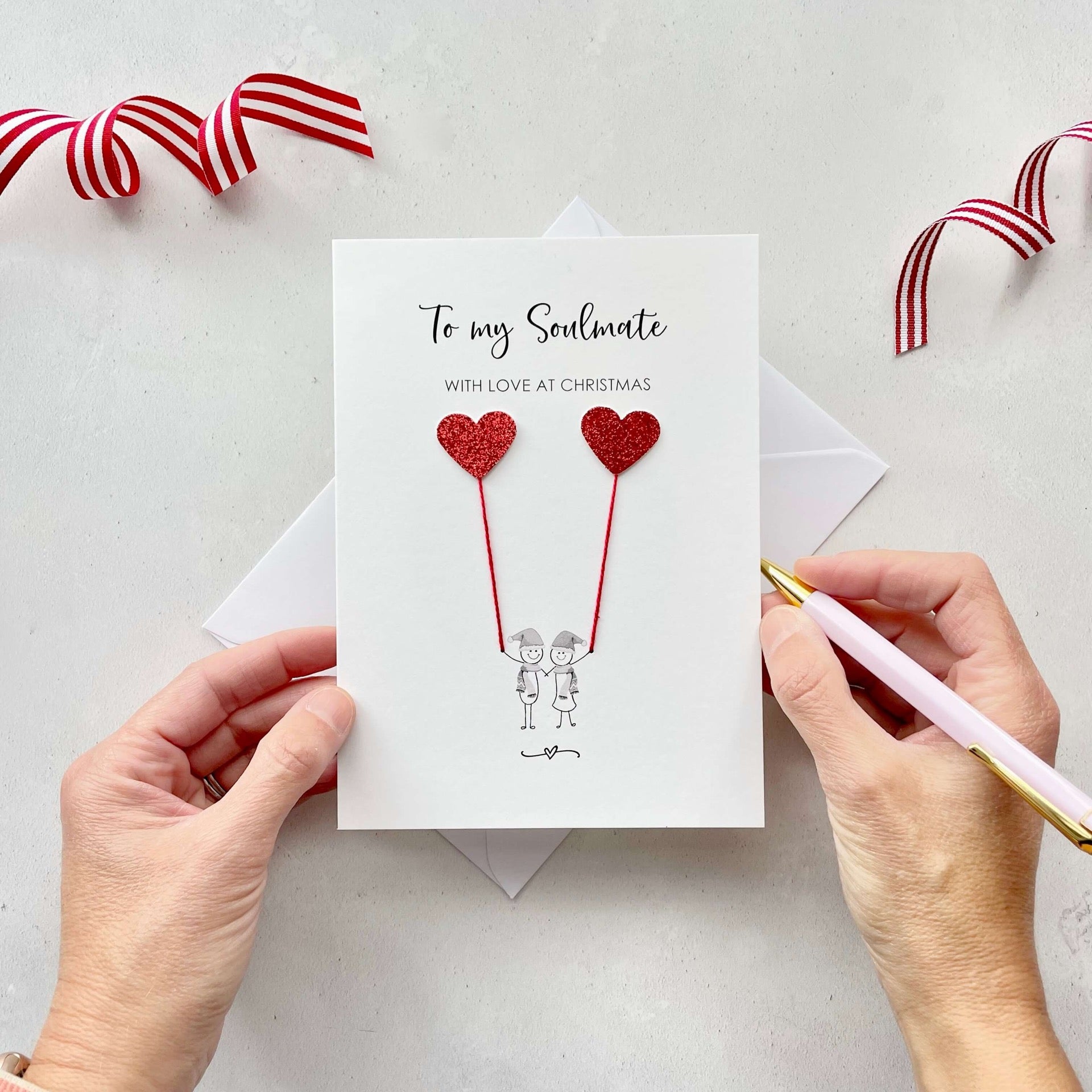 A Christmas card featuring a minimalist design with two figures wearing santa hats holding red glitter heart balloons. The text at the top reads: ‘To my Soulmate, with love at Christmas’. The card is placed on a white background with a white envelope partially visible behind it. A hand is seen holding the card, while another hand holds a pink pen. A red and white striped ribbon is positioned in the top corners of the image for decoration.